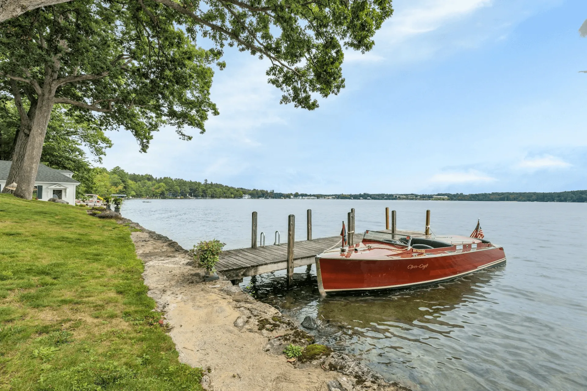 A vintage red boat is docked at a wooden pier beside a lush green lawn, overlooking a serene lake under a clear blue sky.