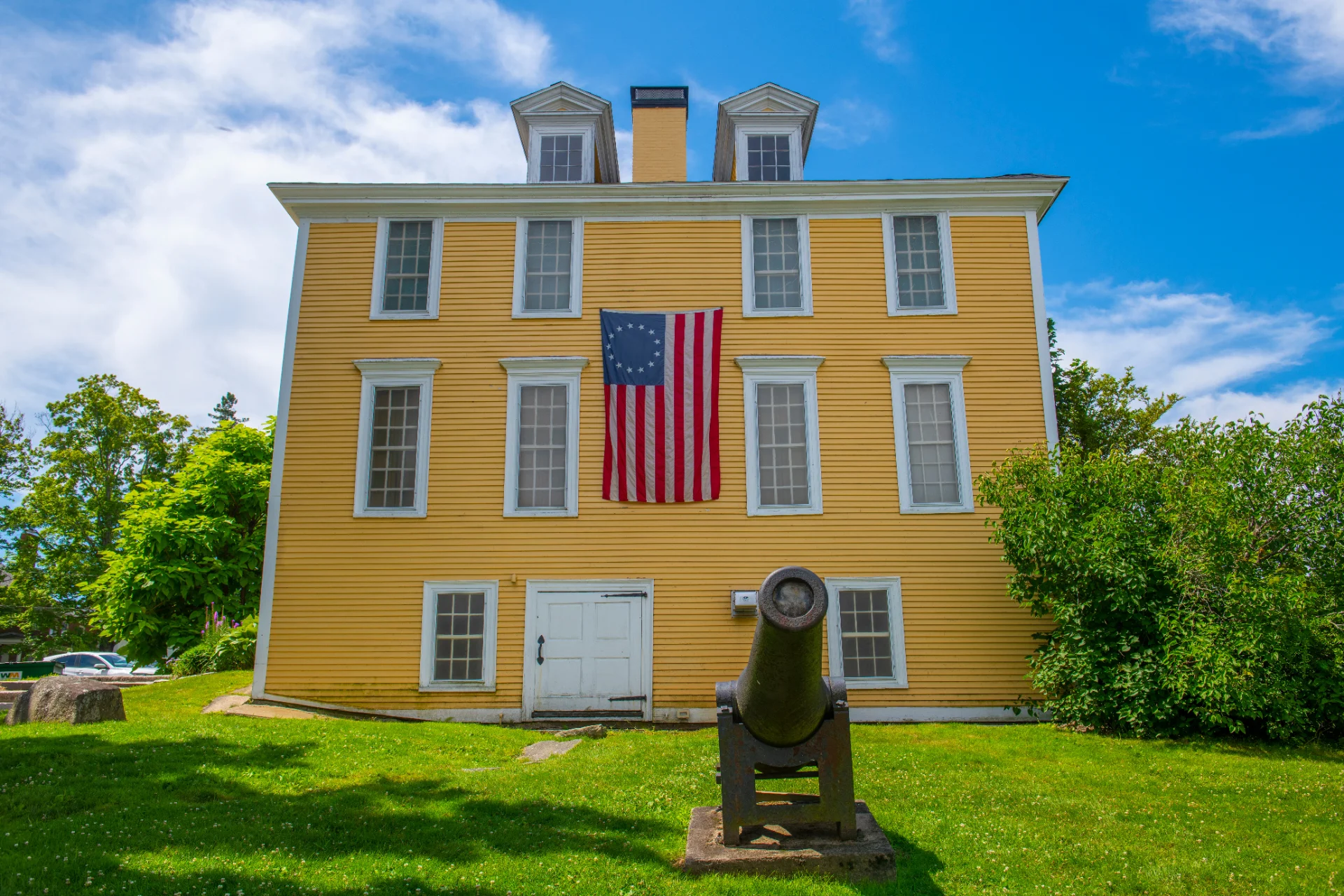 Yellow colonial meeting house with a 13-star flag and a cannon on the lawn