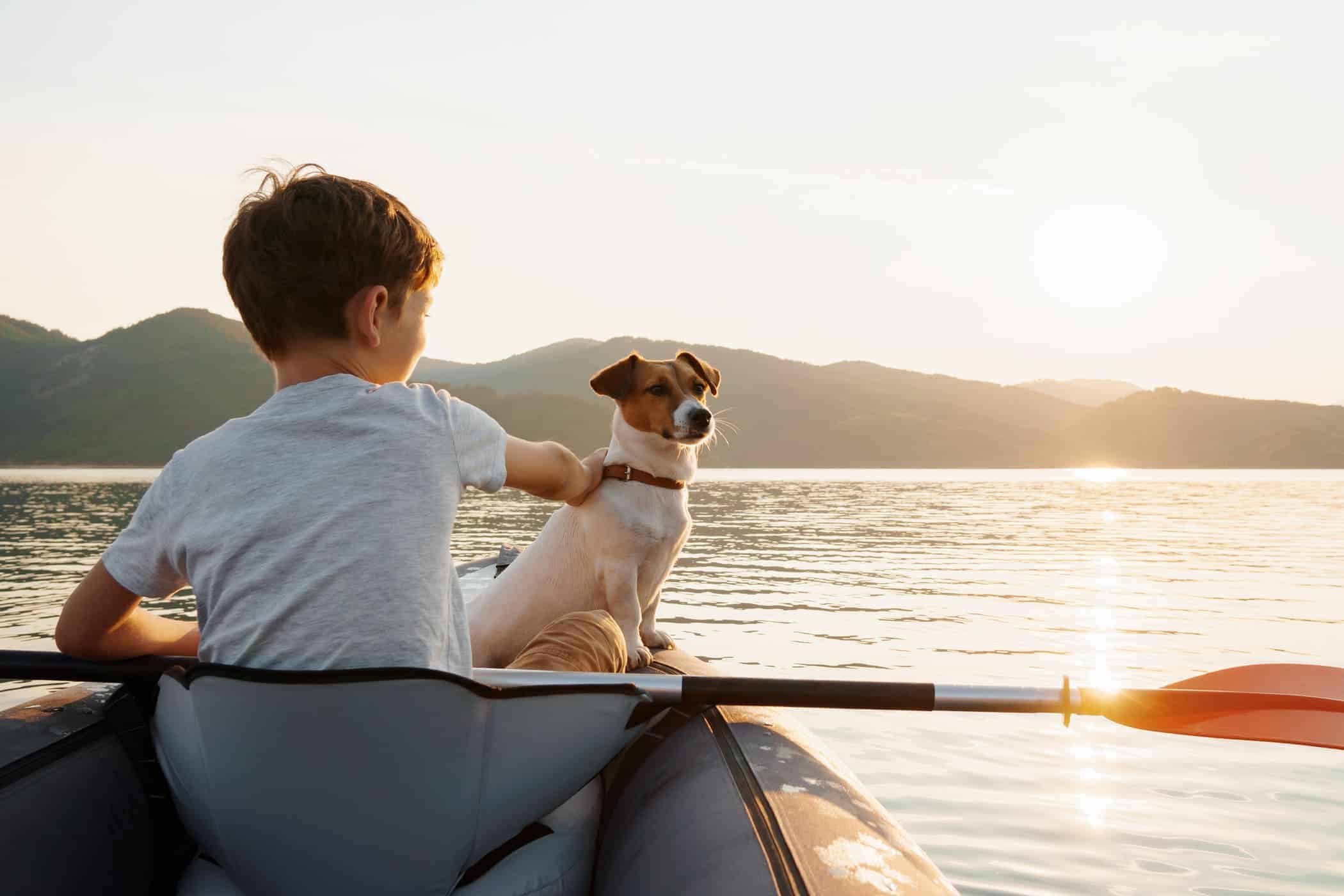 Happy boy with his dog Jack Russell Terrier paddling an inflatable kayak on the water mountain lake against the backdrop of beautiful orange sunset. Family sports vacation. Lens flare. Pet